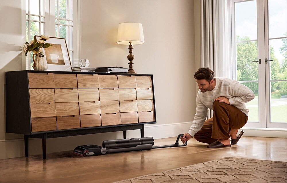 Mężczyzna klęczy, używając odkurzacza. Jest drewniana komoda, okno i lampa.