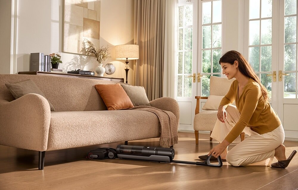 Kobieta odkurza pokój. Beżowa kanapa, lampa i okno w tle. Odkurzacz jest na podłodze.