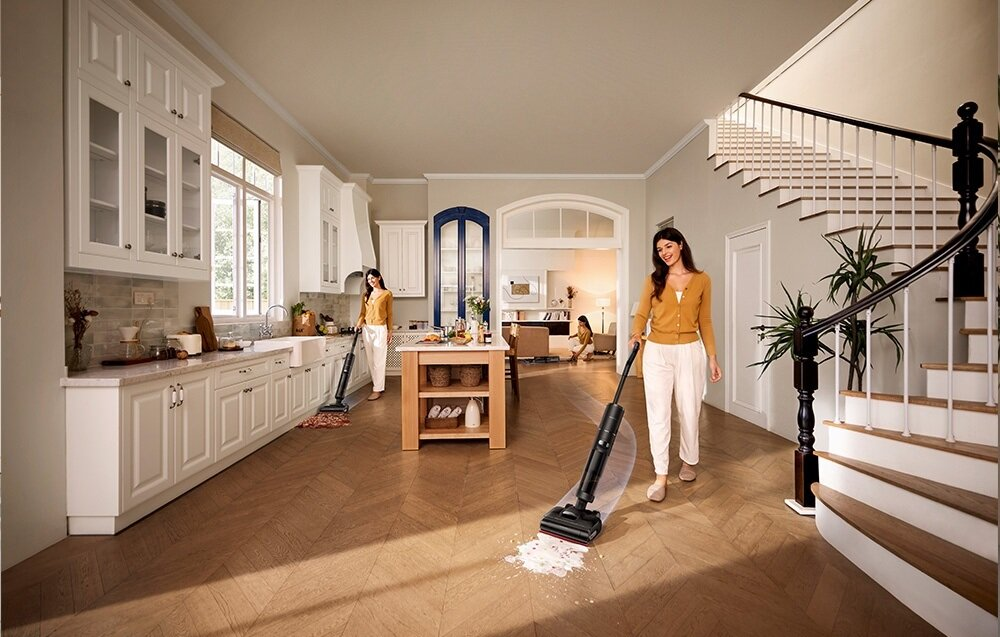 Kobieta odkurza drewnianą podłogę w kuchni. Inna kobieta odkurza w tle. Trzecia kobieta siedzi w tle.