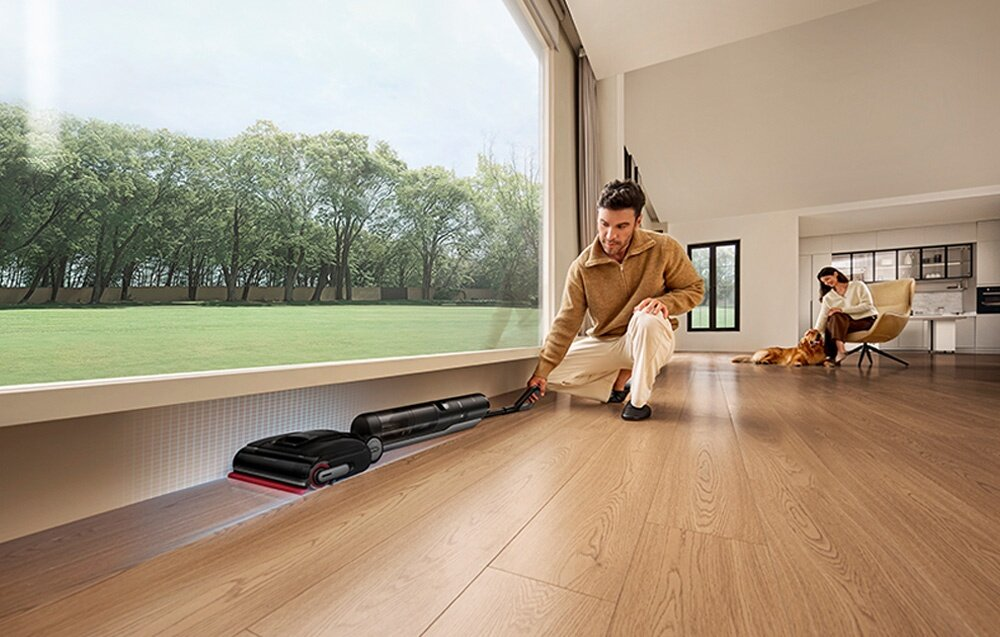 Mężczyzna czyści podłogę odkurzaczem. Kobieta z psem. Na zewnątrz widoczne drzewa i zielona trawa.
