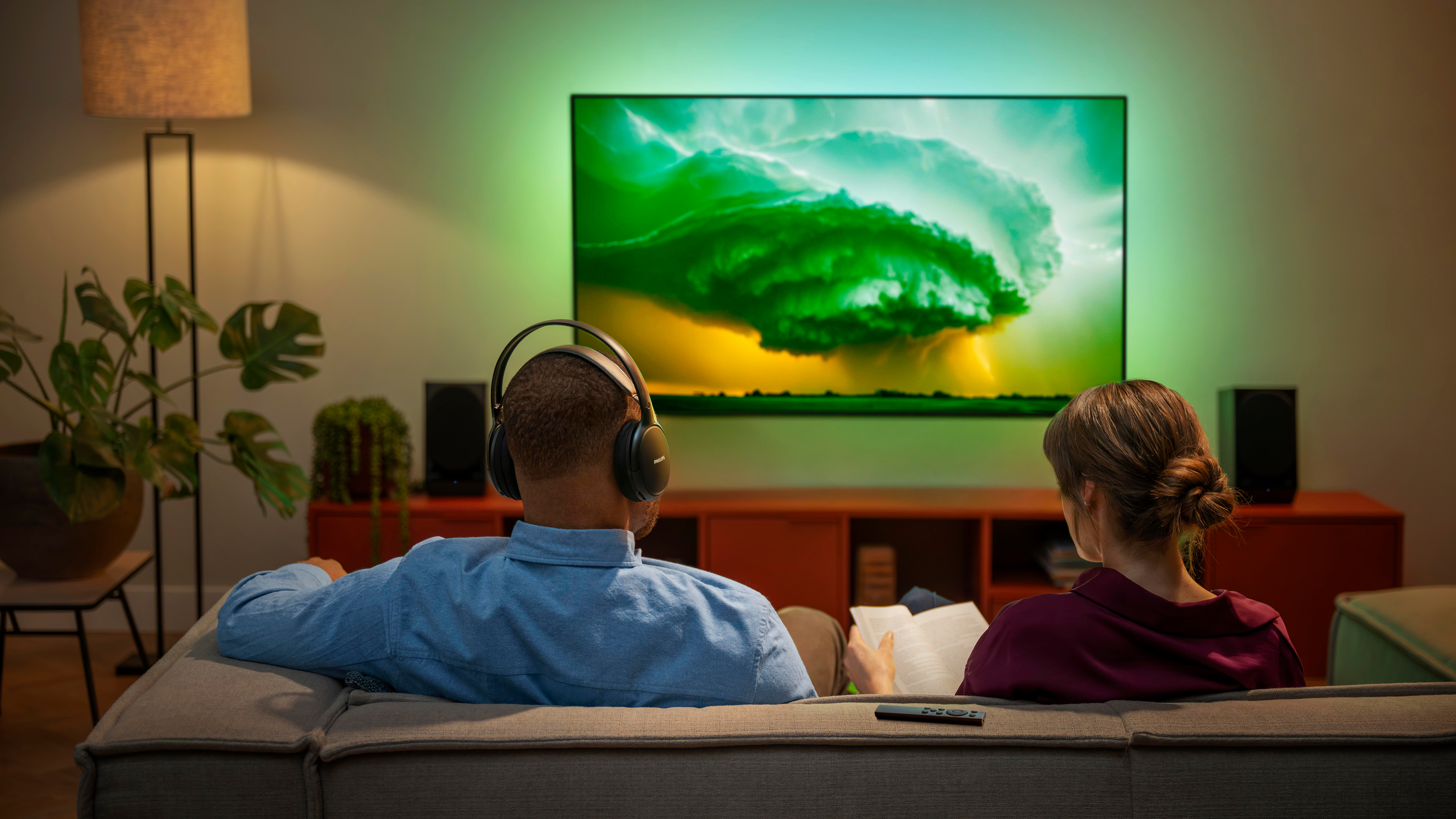 Paar schaut fern, Mann mit Kopfhörern, Frau liest. TV zeigt grüne Sturmszene. Modernes Wohnzimmer.
