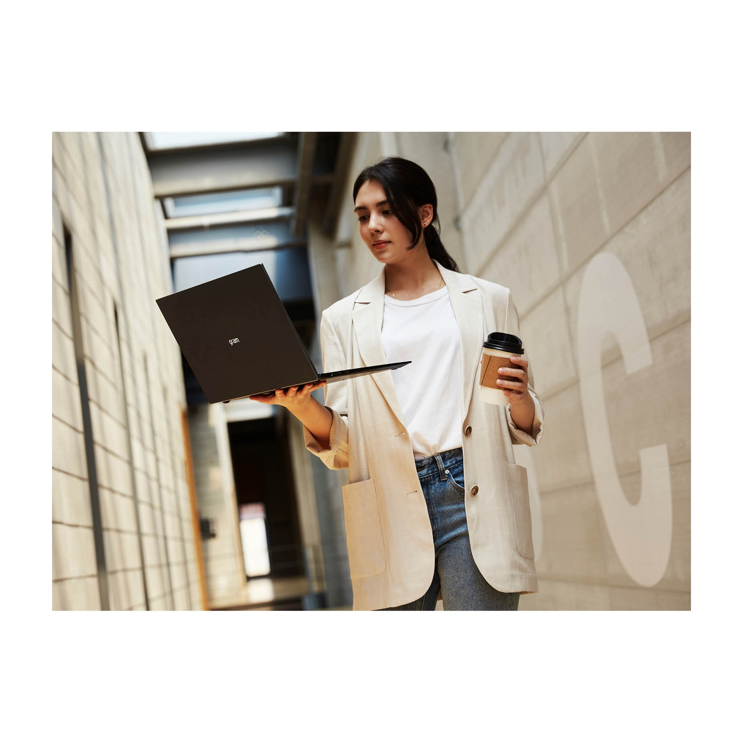 Una donna in giacca tiene in mano un laptop e un caffè. È vicino a un muro di cemento con una grande 'C'.
