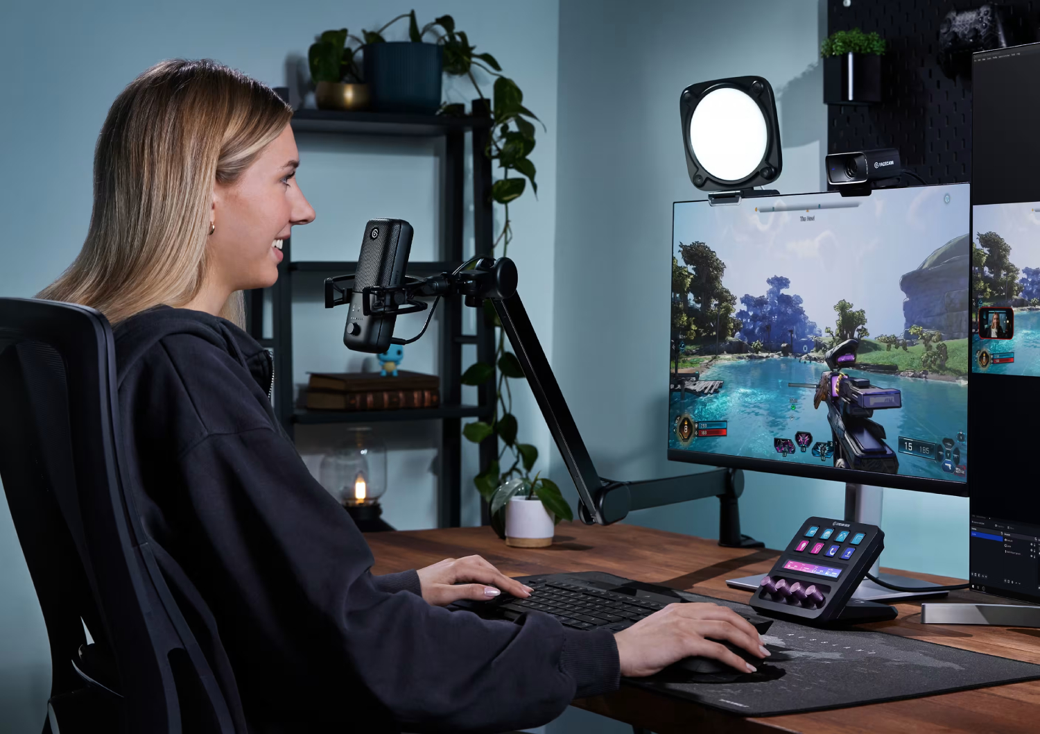 Una mujer sonríe mientras transmite un videojuego. Está en un escritorio con un monitor, micrófono y otros equipos de transmisión.