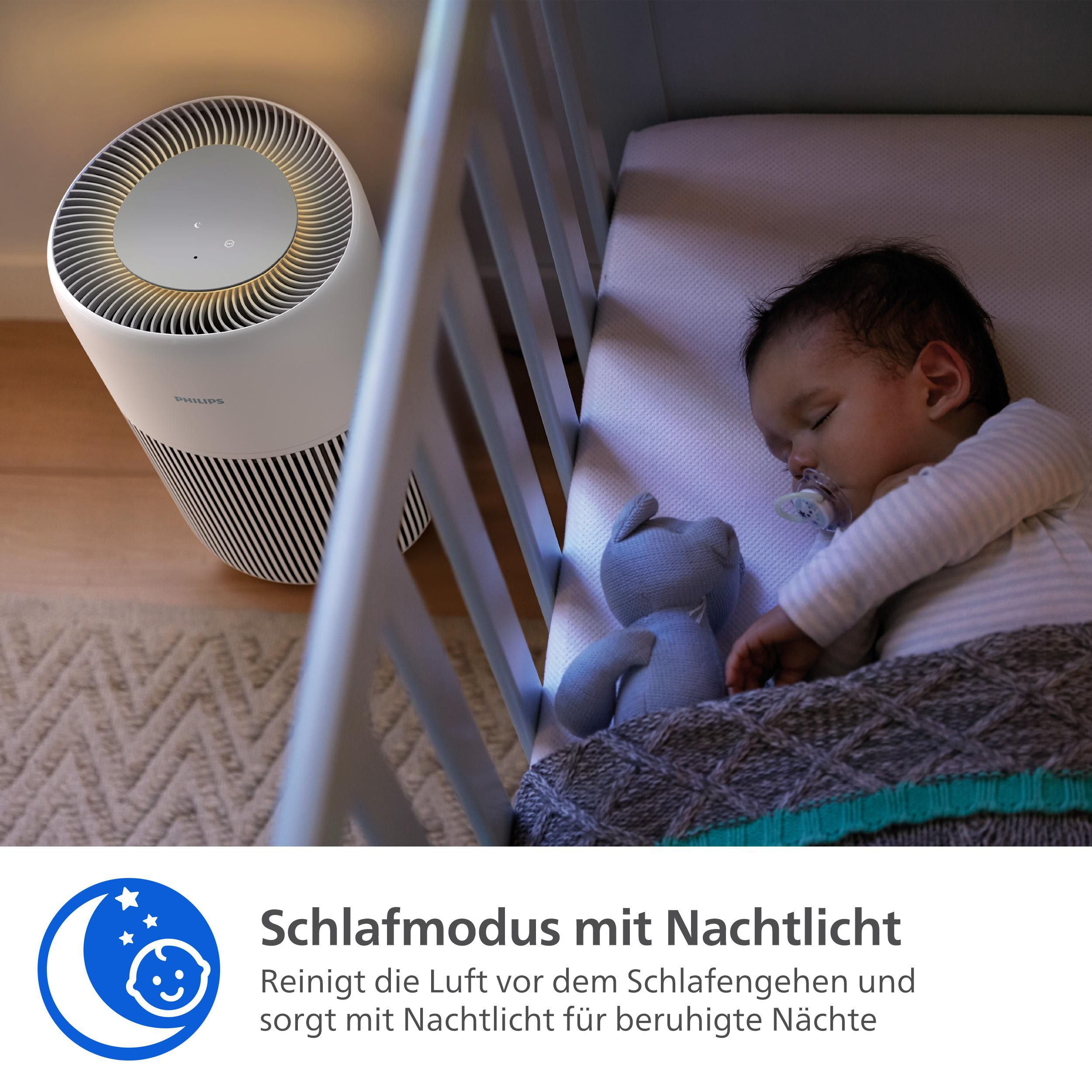 Ein Baby schläft in einem Kinderbett neben einem Luftreiniger. Ein blaues Stofftier liegt neben dem Baby.