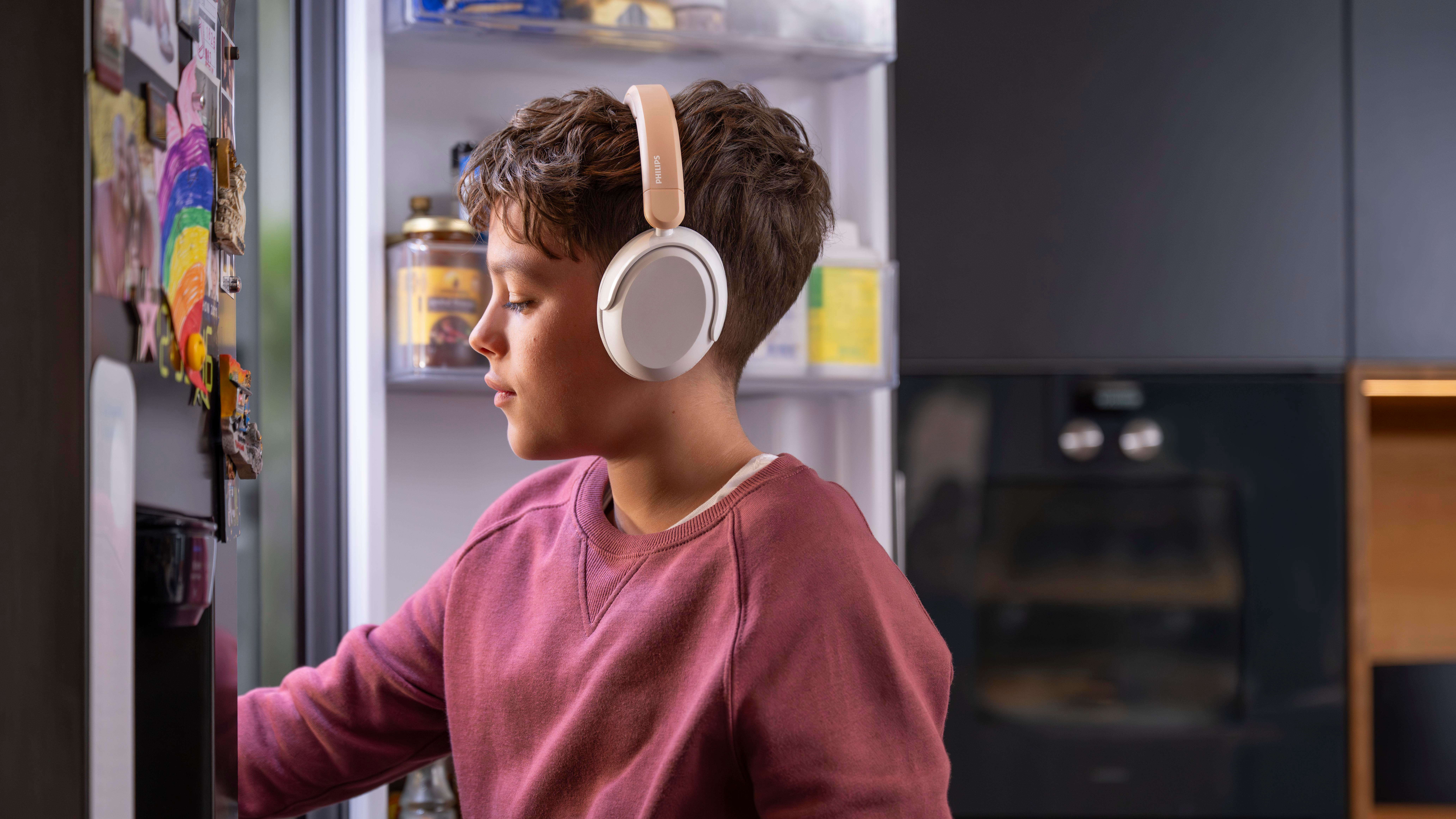 Ein Junge mit Kopfhörern öffnet einen Kühlschrank. Der Kühlschrank ist mit Essen gefüllt. Er trägt einen rosafarbenen Pullover.
