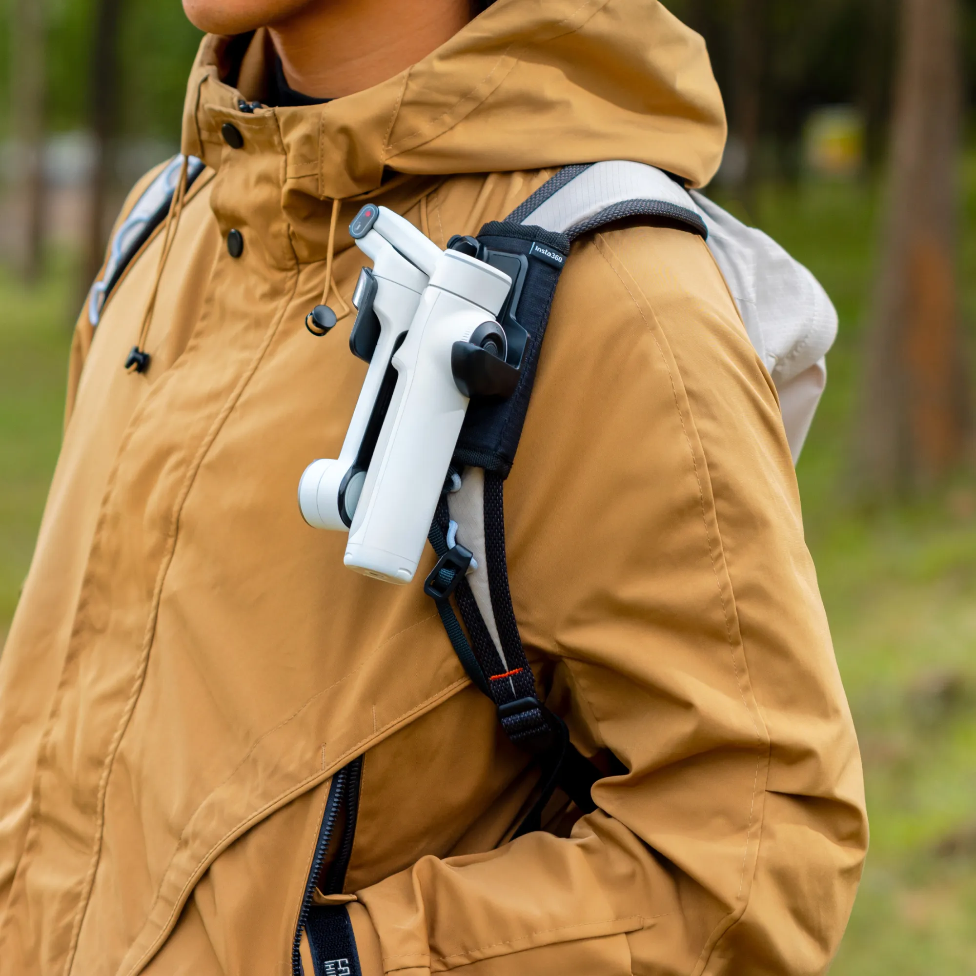 Person mit brauner Jacke und weißer Kamera an einem Rucksackgurt. Grüner Hintergrund.