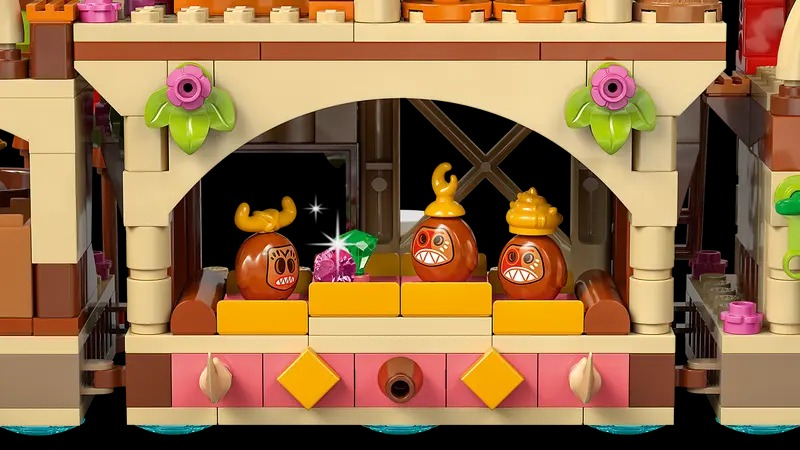 Lego scene with three brown figures with faces, gems, and pink and green decorations. The background is a dark interior.