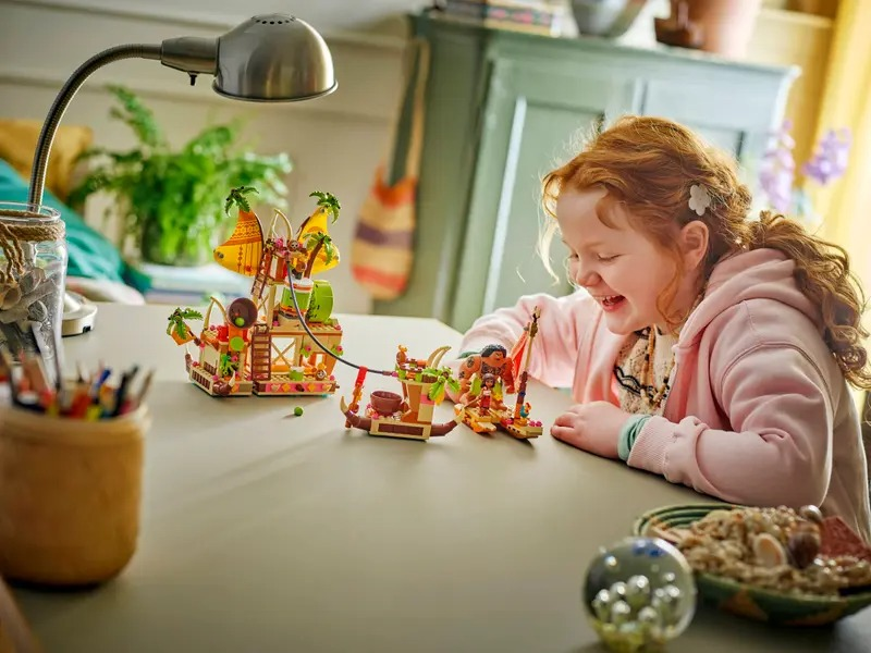 A girl laughs while playing with a Lego set. The set depicts a colorful structure with palm trees, on a table. A lamp is above.