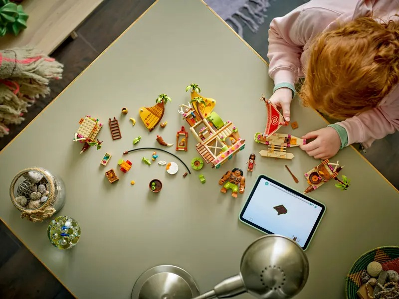 A child with red hair plays with Lego. Lego pieces are scattered on a table. A tablet is on the table showing instructions.