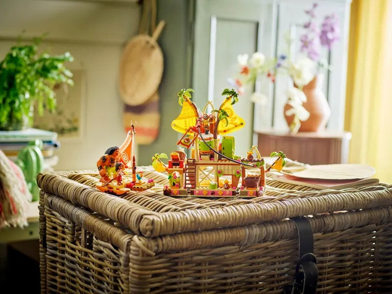 A Lego set of a tropical island scene sits on a wicker chest. Figures include Maui and Moana. Other elements include a boat and a hut.