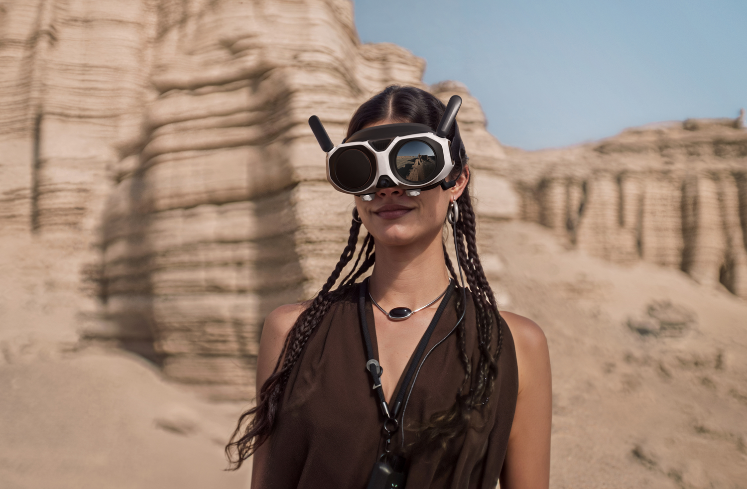 Una mujer usa gafas de realidad virtual, de pie en un paisaje desértico. El fondo tiene formaciones rocosas en capas. Tiene trenzas y una parte superior oscura.
