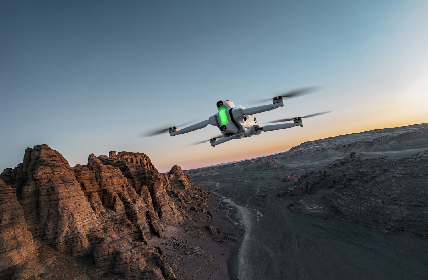 Un dron vuela sobre un cañón. El dron es blanco con luces verdes. El cañón es marrón y gris. El cielo es azul y naranja.