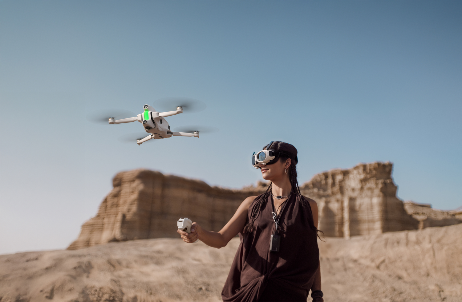 Una mujer controla un dron en el desierto. El dron flota sobre ella. Lleva gafas y un vestido oscuro.