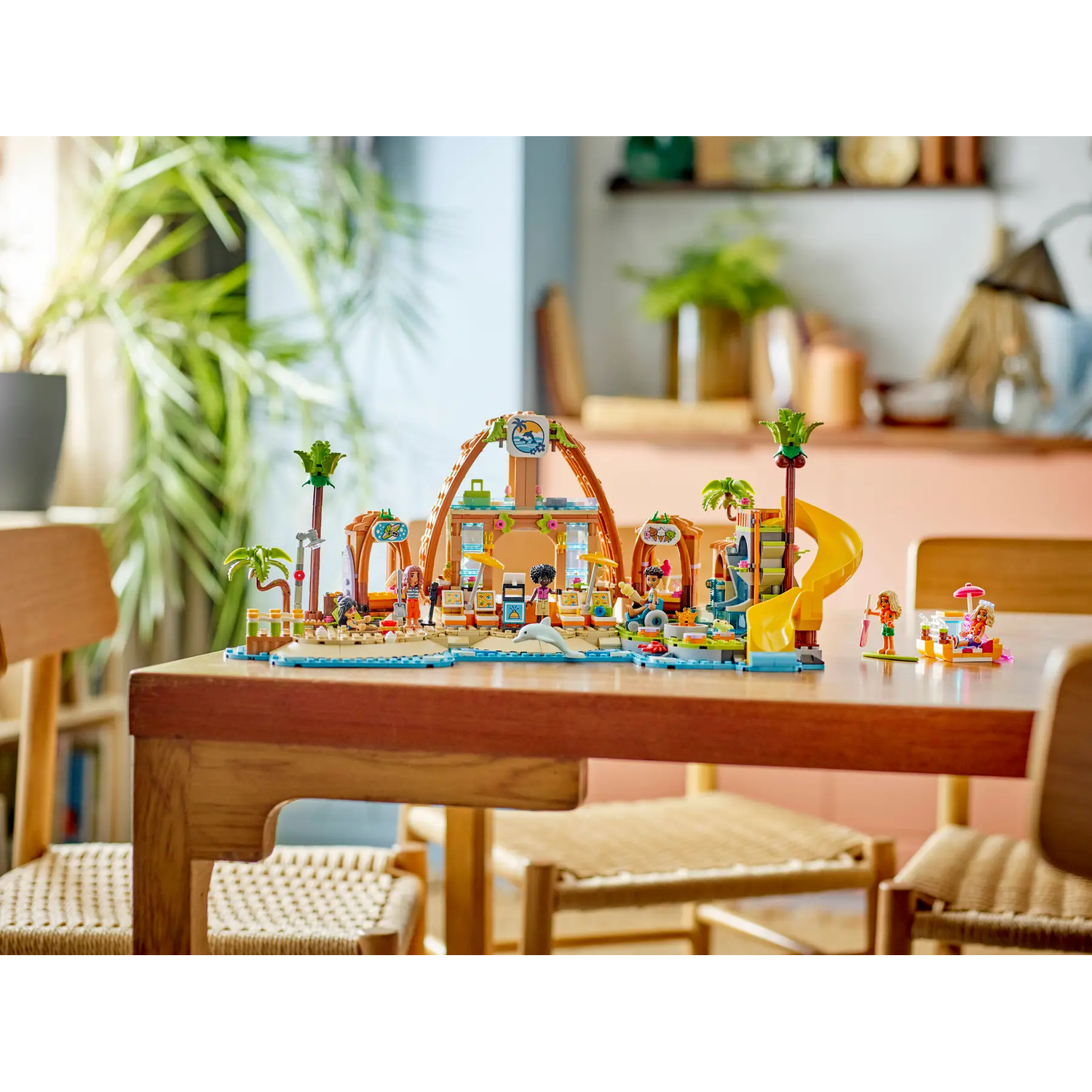 A Lego beach scene with palm trees, a slide, and figures on a wooden table. Background shows plants and a room setting.