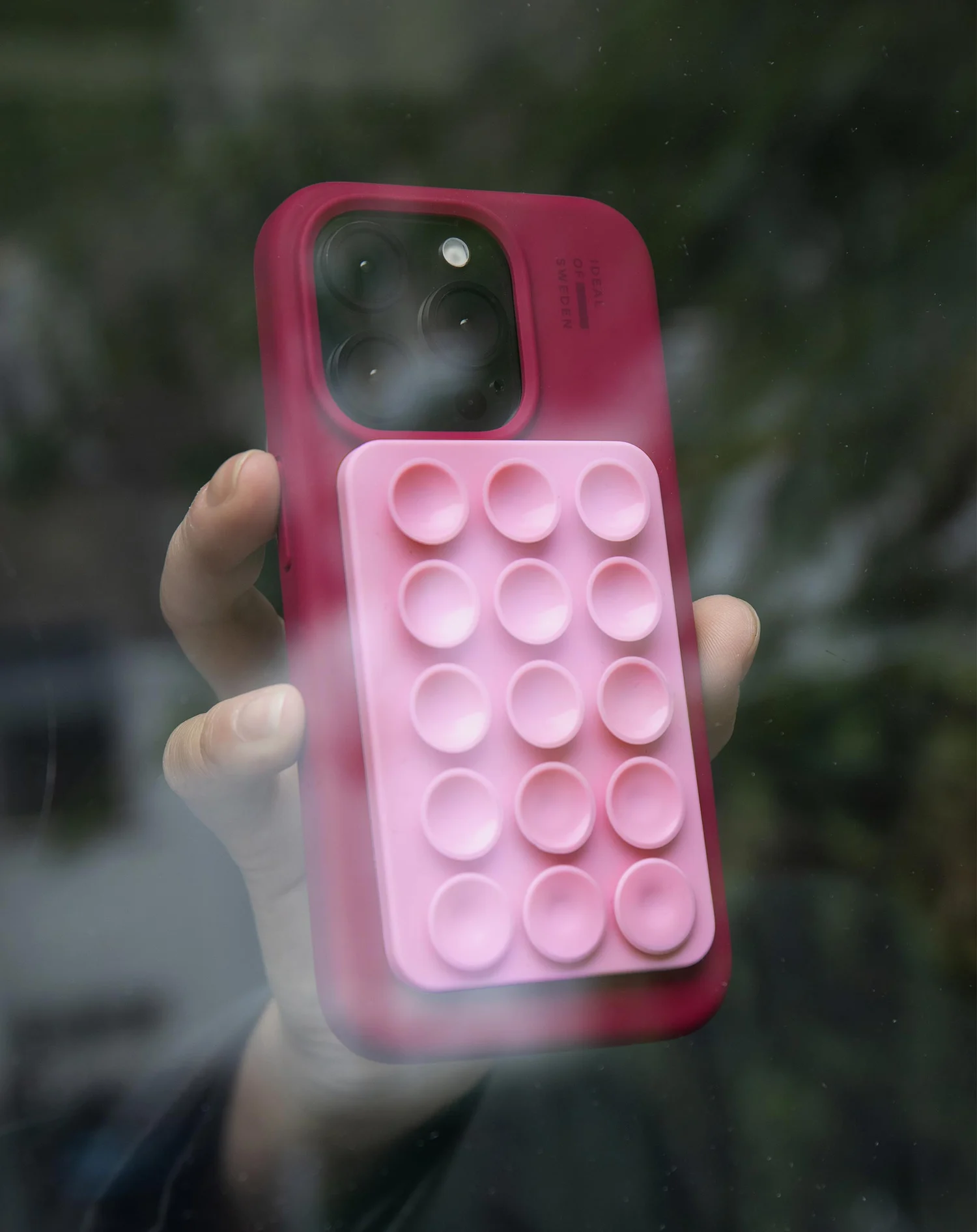 Egy kéz egy rózsaszín telefontokot tart, benne egy buborékpattogtató játékkal. A háttér egy elmosódott zöld kültéri jelenet.