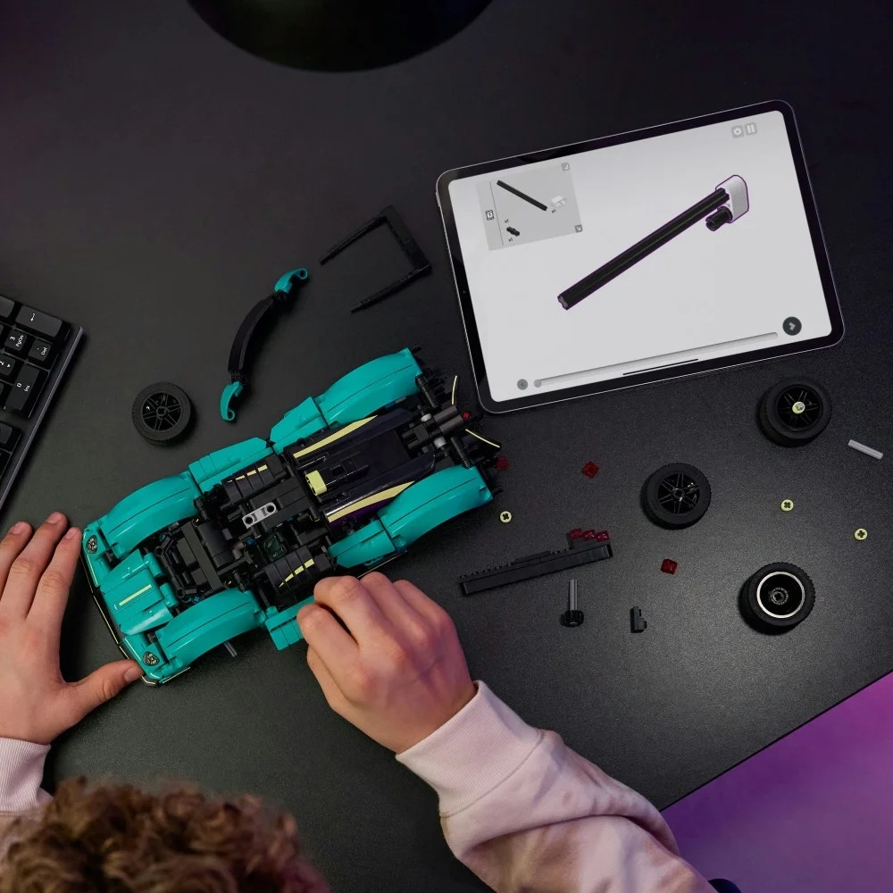 Osoba składająca turkusowo-czarny samochód Lego z instrukcjami na tablecie. Części i koła są rozrzucone na czarnym biurku.