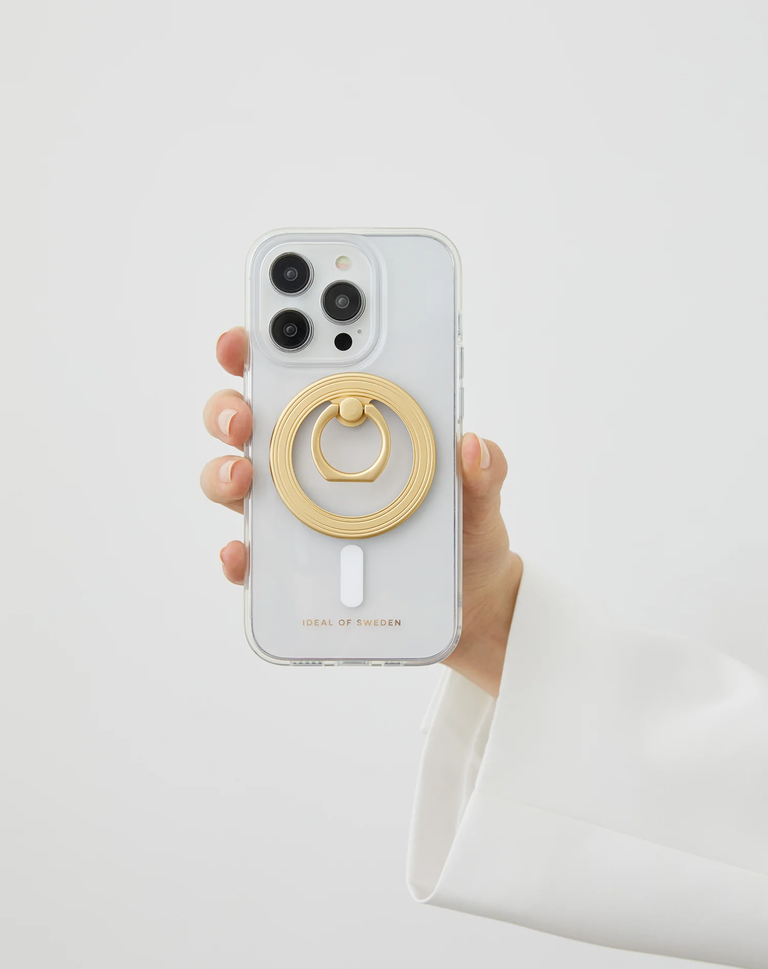 Egy kéz egy telefont tart, rajta egy arany gyűrűtartóval. A telefon átlátszó tokban van. Fehér háttér.
