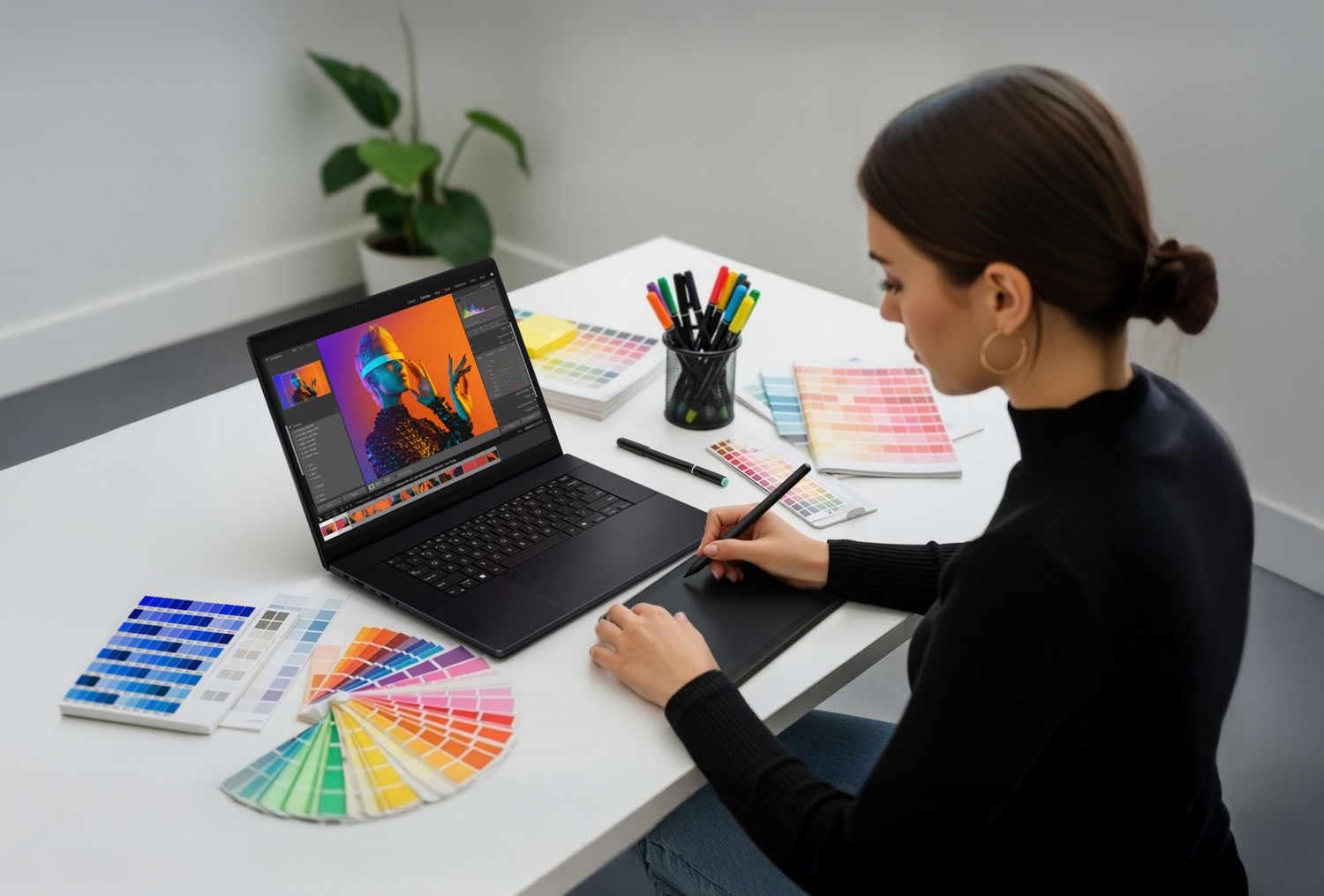 Mujer trabajando en portátil, editando imagen. Paletas de colores y bolígrafos en el escritorio. Planta en el fondo. Lleva camisa negra.