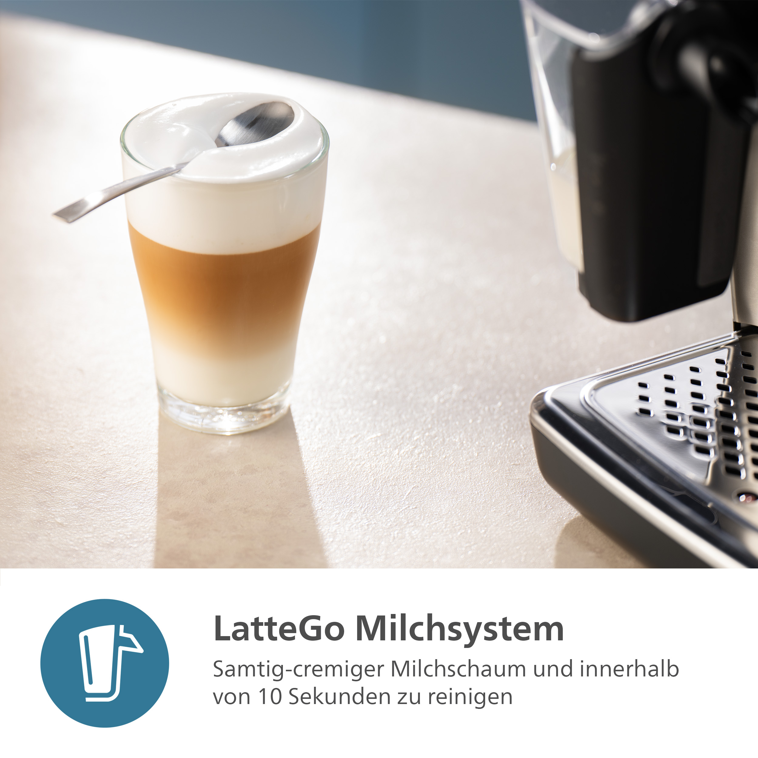 Ein Latte in einem Glas mit einem Löffel. Rechts ist eine Kaffeemaschine zu sehen. Der Hintergrund ist unscharf.