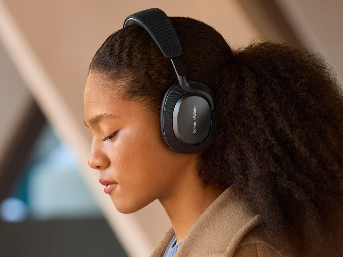 Egy göndör hajú nő fekete fejhallgatót visel, csukott szemmel, egy világos térben. A fejhallgató Bowers & Wilkins.