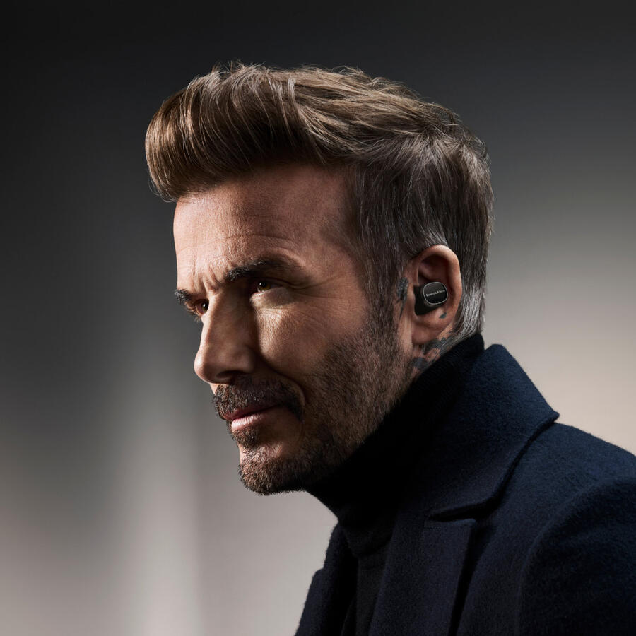 David Beckham fülhallgatót visel, oldalra néz. Fekete garbót és kabátot visel. A háttér szürke.