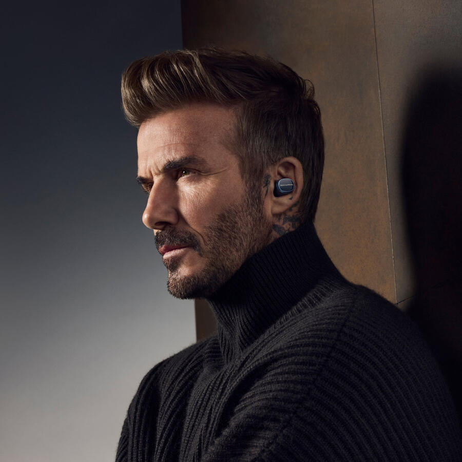 David Beckham fekete garbót és fülhallgatót visel. Komolyan oldalra néz. A háttér barna.