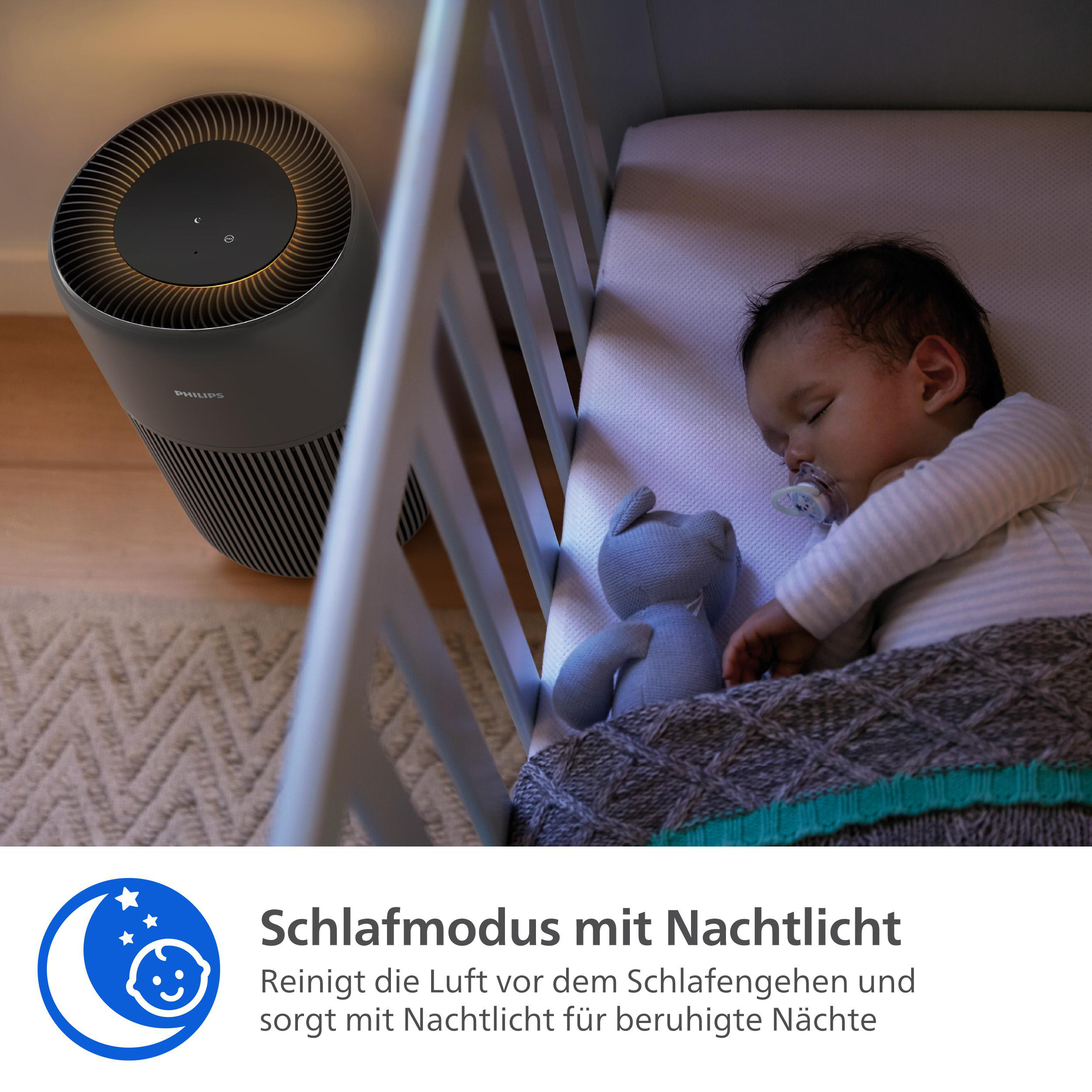 Ein Baby schläft in einem Kinderbett mit einem Stofftier und einer Decke. Im Hintergrund befindet sich ein Luftreiniger.