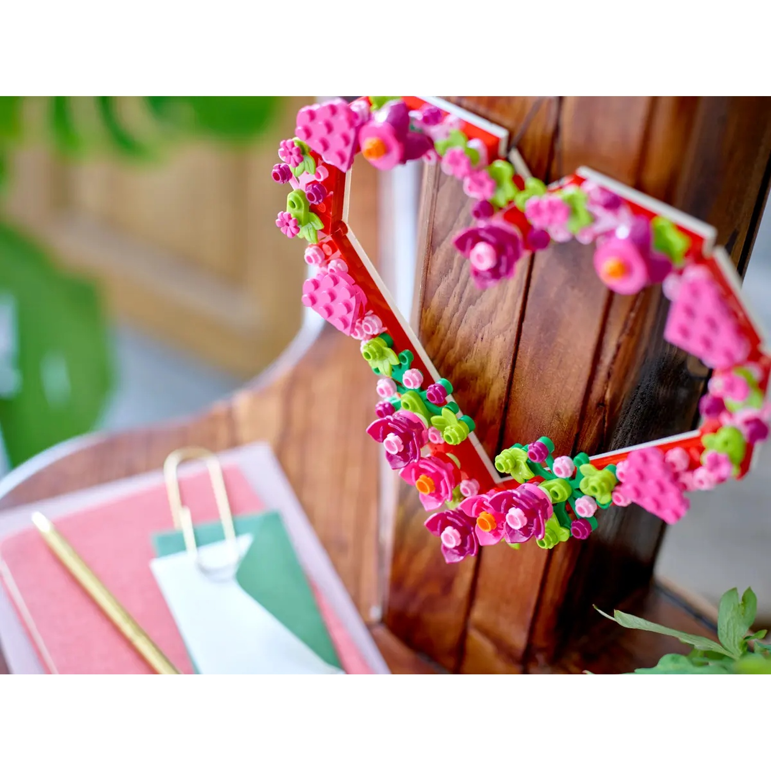 Une décoration en forme de cœur avec des fleurs roses. C'est sur une surface en bois, à côté de papiers roses et verts avec un stylo doré.