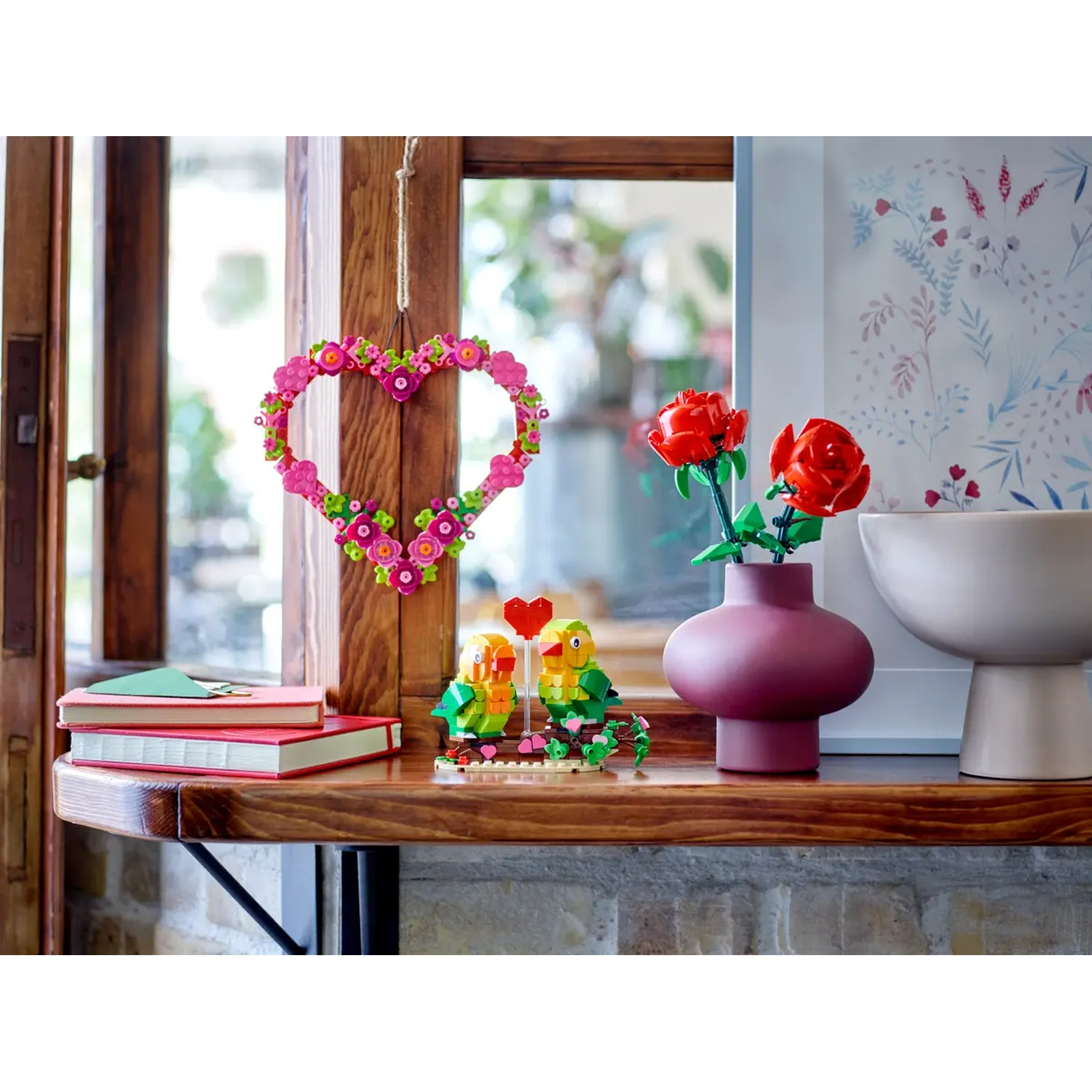 Une étagère en bois présente des fleurs Lego, des oiseaux d'amour, des livres et une couronne en forme de cœur. Une fenêtre avec de la lumière est en arrière-plan.