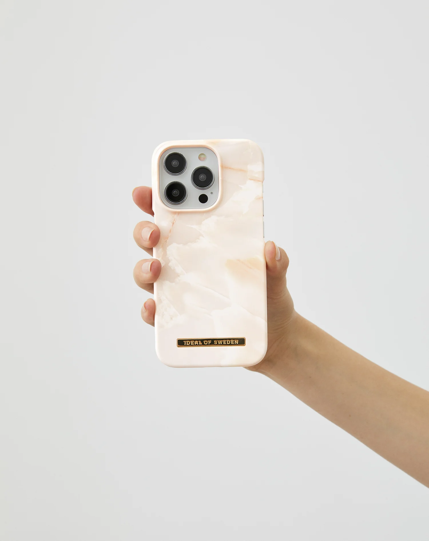 Egy kéz márványmintás telefontokot tart fehér háttér előtt. A tokon egy arany Ideal of Sweden felirat található.