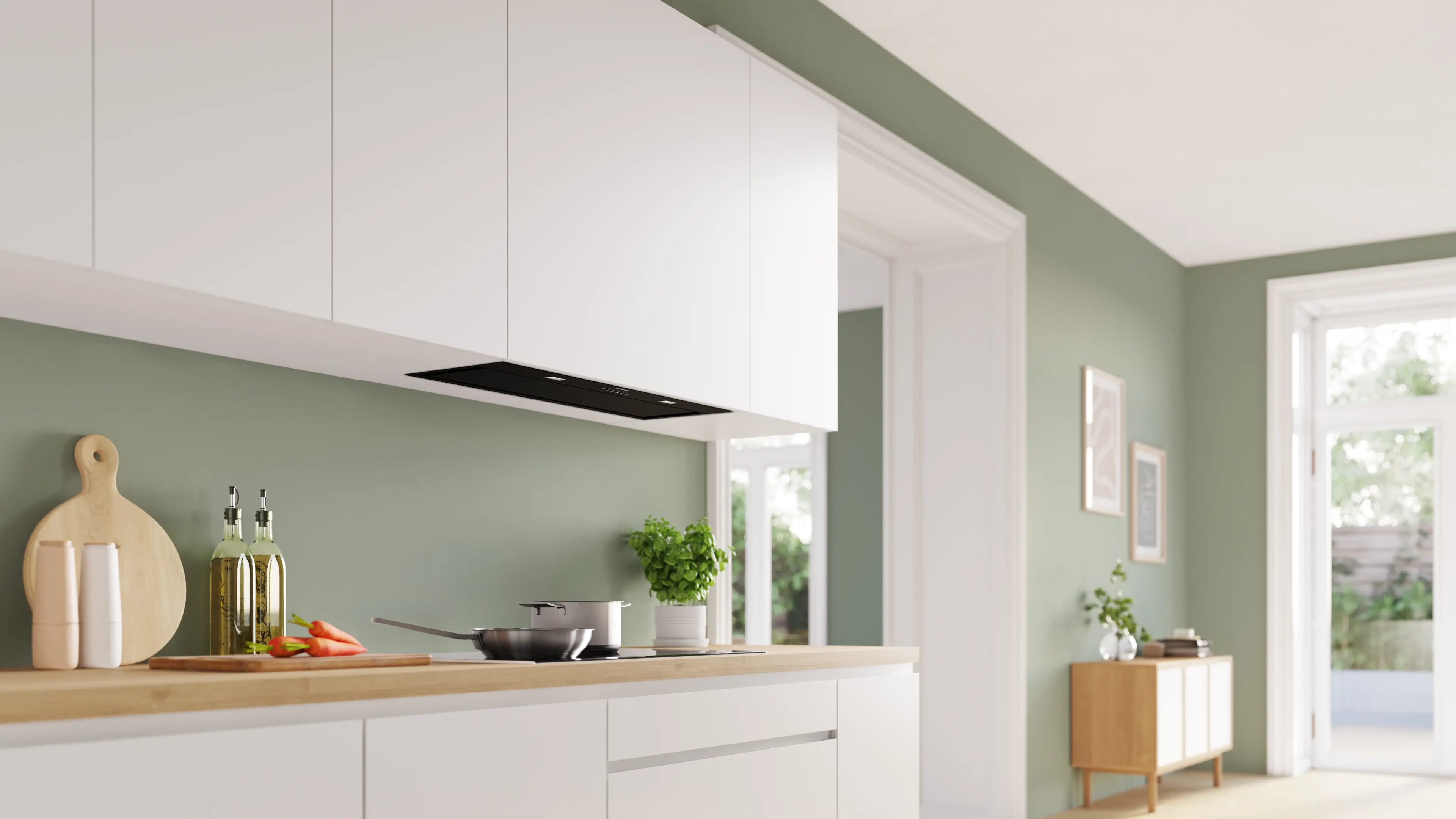 Cocina moderna con armarios blancos, encimera de madera y pared verde oliva. Incluye sartén, zanahorias y albahaca.