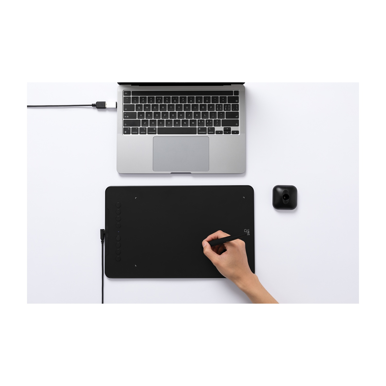 Un laptop, una tavoletta grafica nera con penna e una fotocamera nera sono su una superficie bianca. Una mano usa la penna sulla tavoletta.