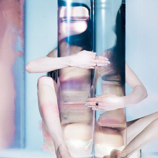 Eine Person in einer transparenten Röhre, Ringe an den Fingern. Der Hintergrund hat rosa und blaue Farbtöne.