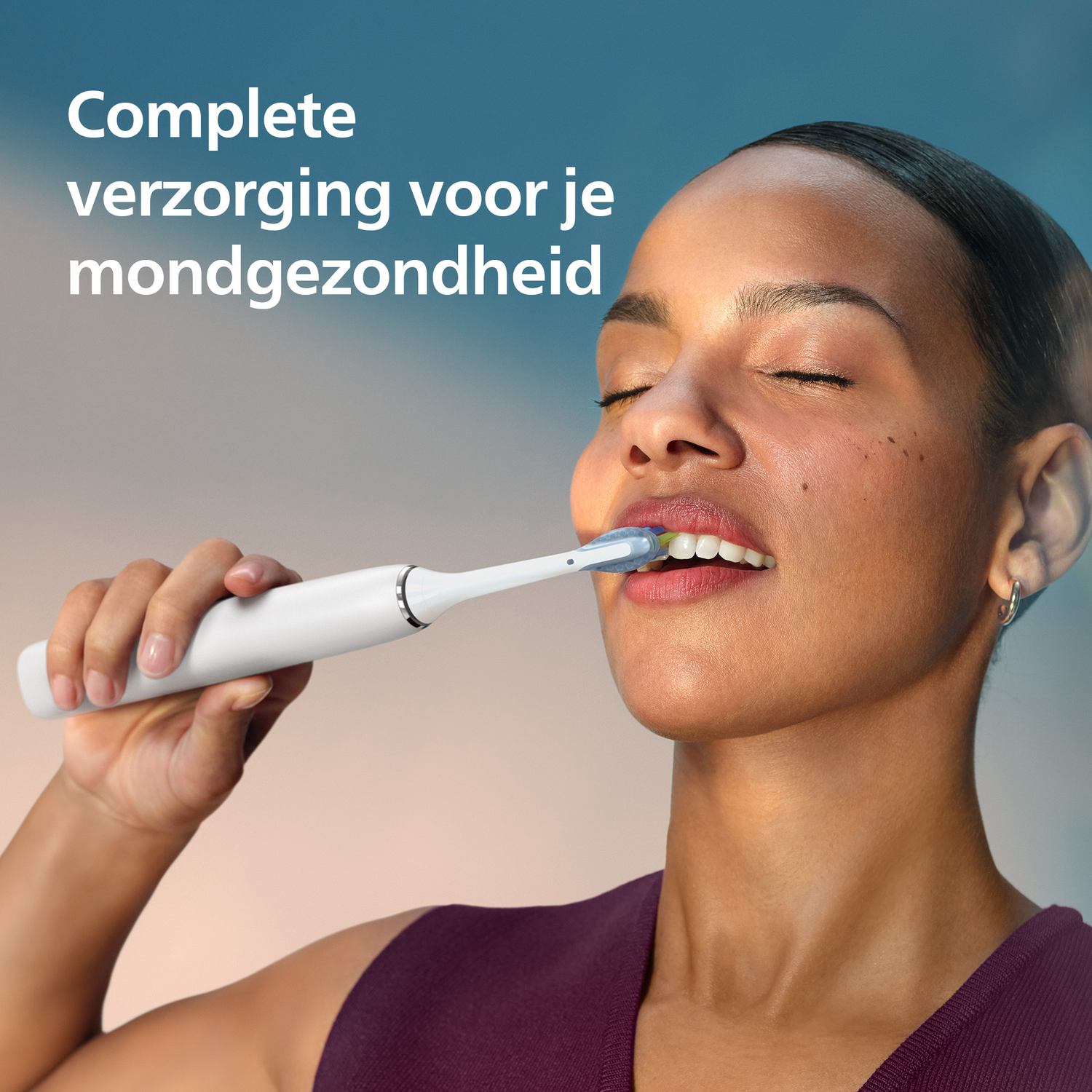 Een vrouw poetst haar tanden met een elektrische tandenborstel. Ze heeft een donkere huid, gesloten ogen en een bordeauxrode top. Tekst in het Nederlands zichtbaar.