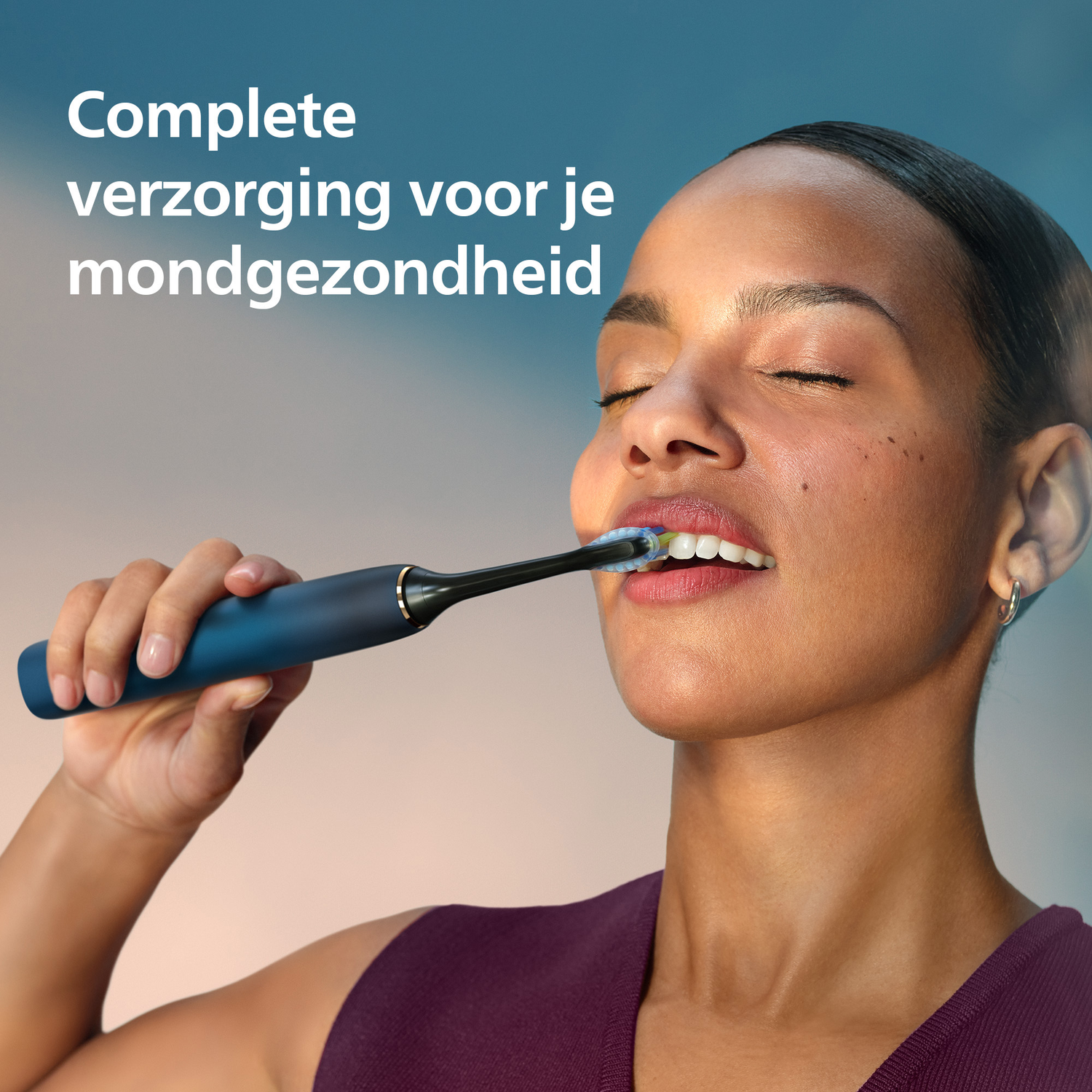 A woman with closed eyes brushes her teeth with a blue electric toothbrush. Text above reads: Complete verzorging voor je mondgezondheid.