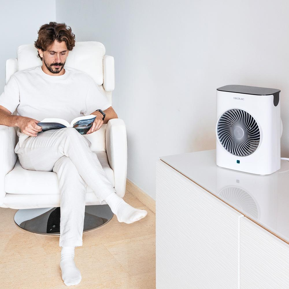 Un hombre lee un libro en una silla blanca. Un ventilador blanco está sobre una mesa blanca. La habitación es mayormente blanca.