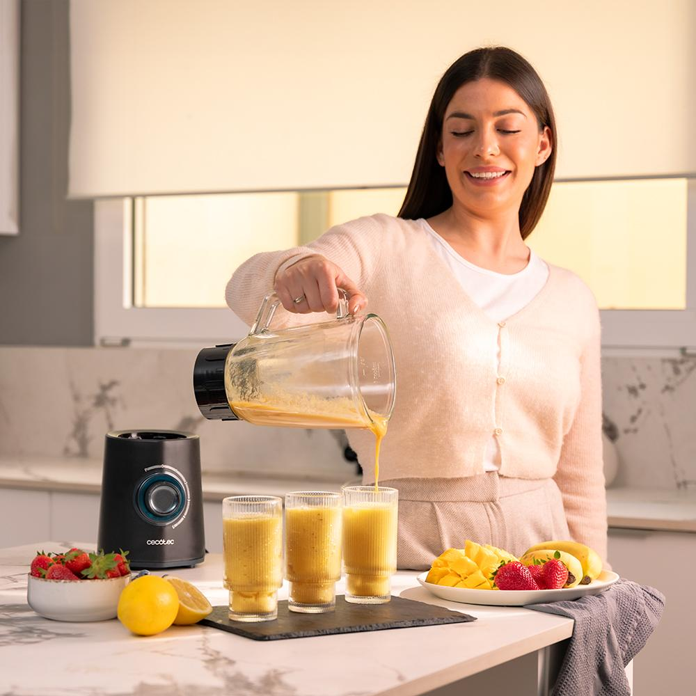 Mujer vertiendo batido en vasos. Licuadora, fruta y un plato de comida en la encimera. La mujer sonríe.