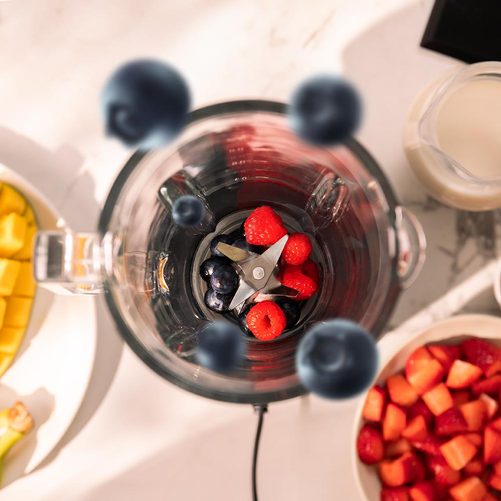Una licuadora llena de arándanos y frambuesas, rodeada de ingredientes como mango, fresas y una jarra de líquido sobre una superficie blanca.