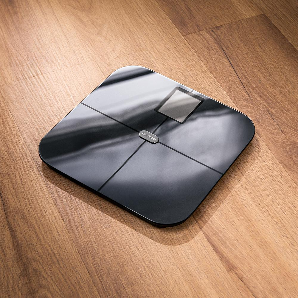 Una báscula digital negra sobre un suelo de madera. La báscula tiene una pantalla y es visible la marca Cecotec.