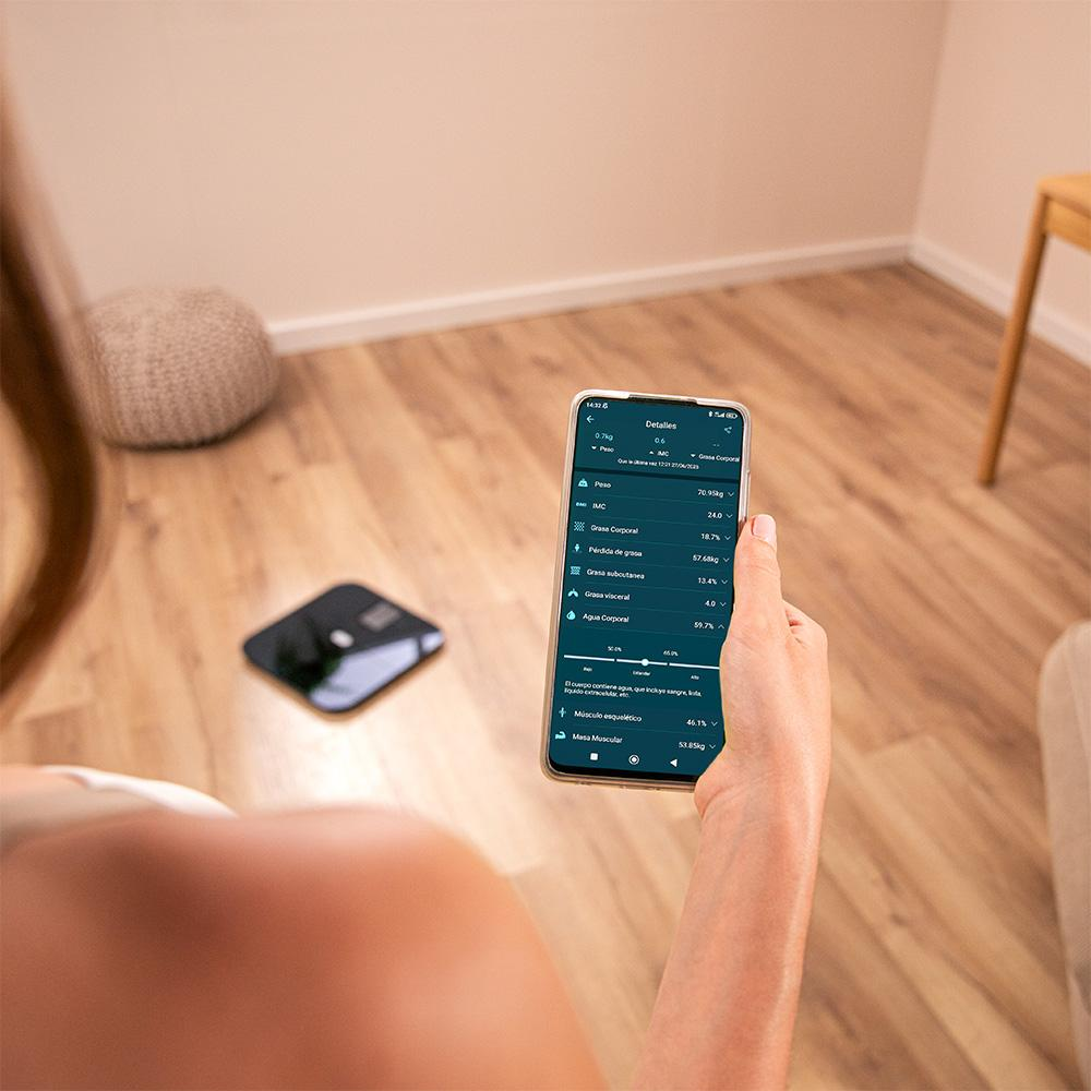Una persona sostiene un teléfono que muestra detalles de la composición corporal. Una báscula está en el suelo de madera. Un puf beige está en el fondo.