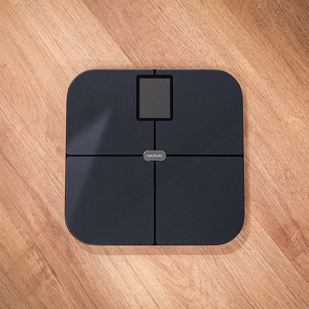 Una báscula digital negra sobre un suelo de madera. Tiene una pantalla y la marca 'cecotec'. El suelo es de madera marrón claro.