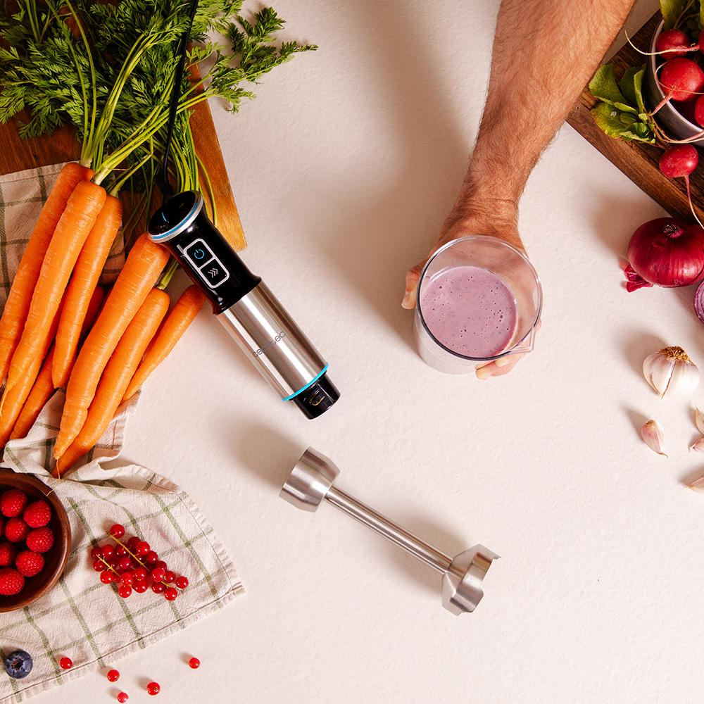Una persona sostiene un vaso de batido morado. Zanahorias, batidora y verduras están sobre la mesa. Se ven bayas y cebolla roja.