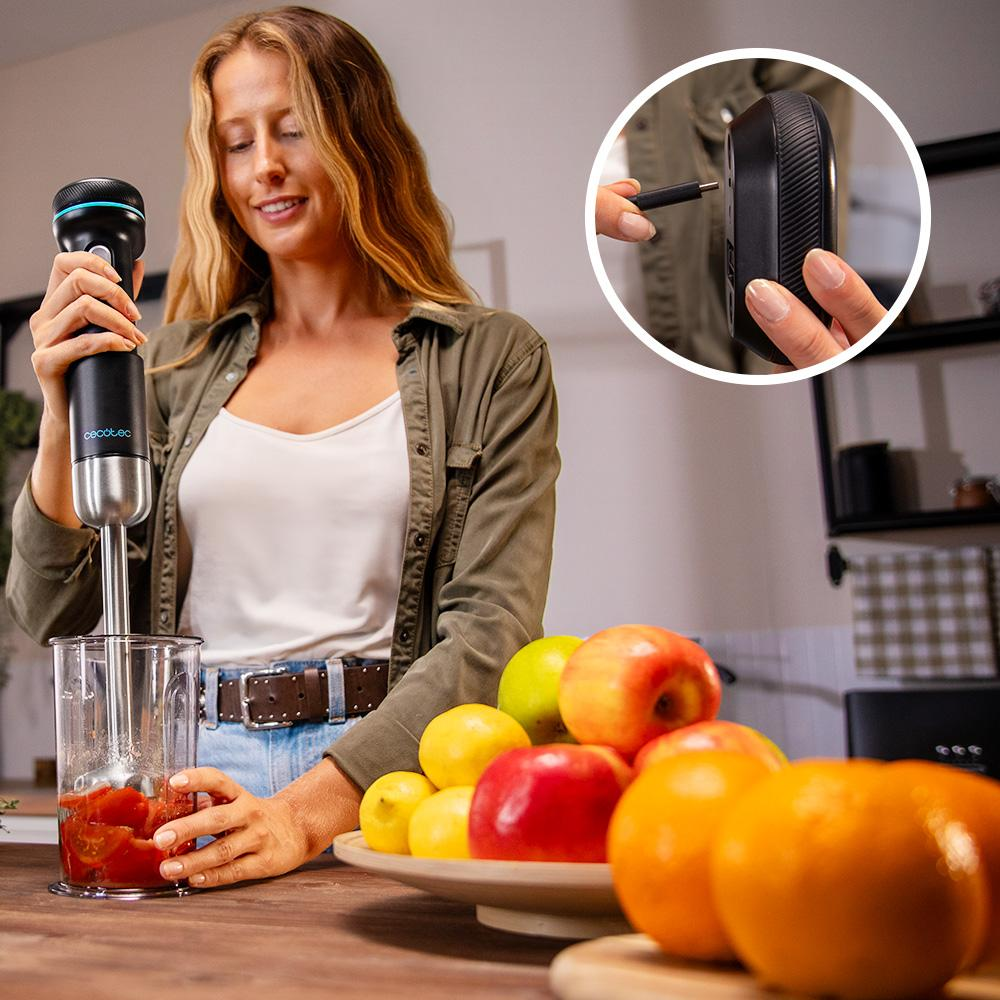 Mujer mezclando tomates con una batidora. Frutas en un plato delante. Otra mano enchufa un cable negro a un dispositivo.