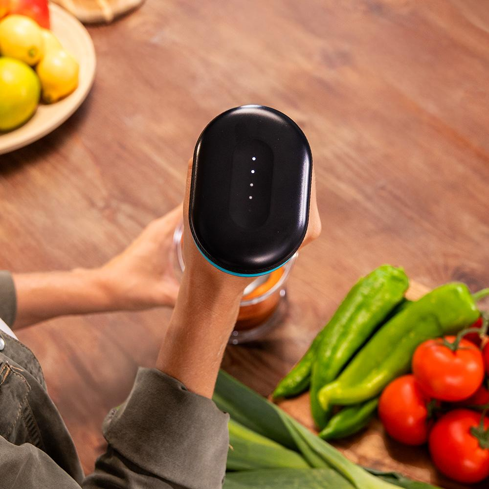 Una persona sostiene un electrodoméstico de cocina negro. Verduras, incluidos tomates y pimientos, sobre una superficie de madera.