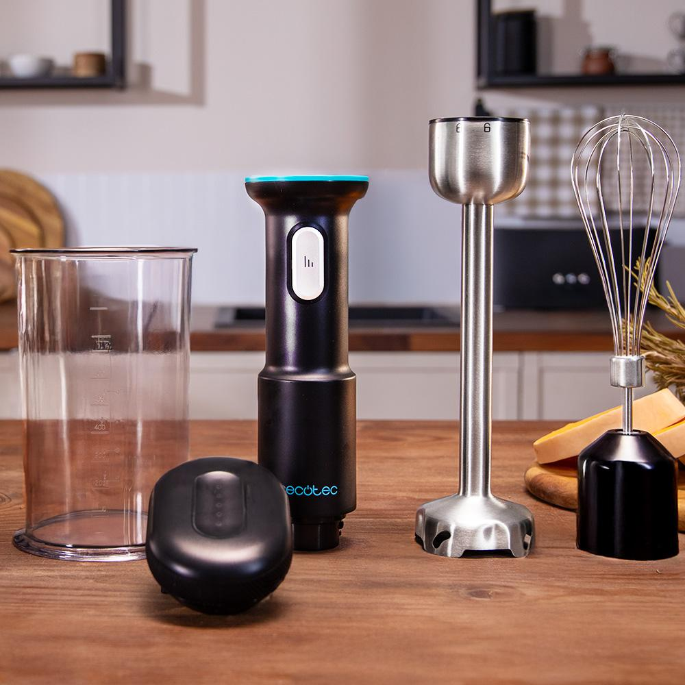Una mesa de madera exhibe un juego de licuadora: vaso, base negra, batidora de inmersión, batidor y un cuerpo de licuadora negro. Fondo: cocina.