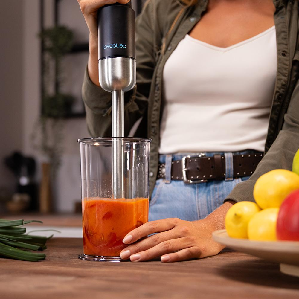 Una persona usa una licuadora en un vaso para mezclar líquido naranja. Hay frutas a la derecha y cebolletas a la izquierda.