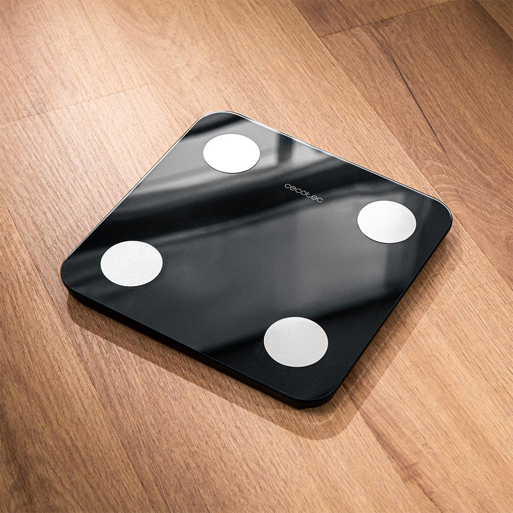 Una báscula de baño digital negra con cuatro círculos blancos sobre un suelo de madera. Se ve la marca 'cecotec'.