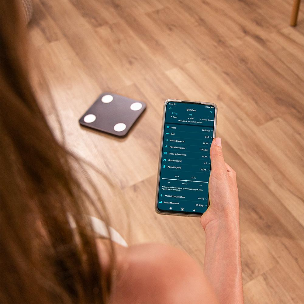 Una persona sostiene un teléfono que muestra detalles de la composición corporal. Una báscula está en el suelo de madera. El teléfono muestra peso, porcentaje de grasa y masa muscular.