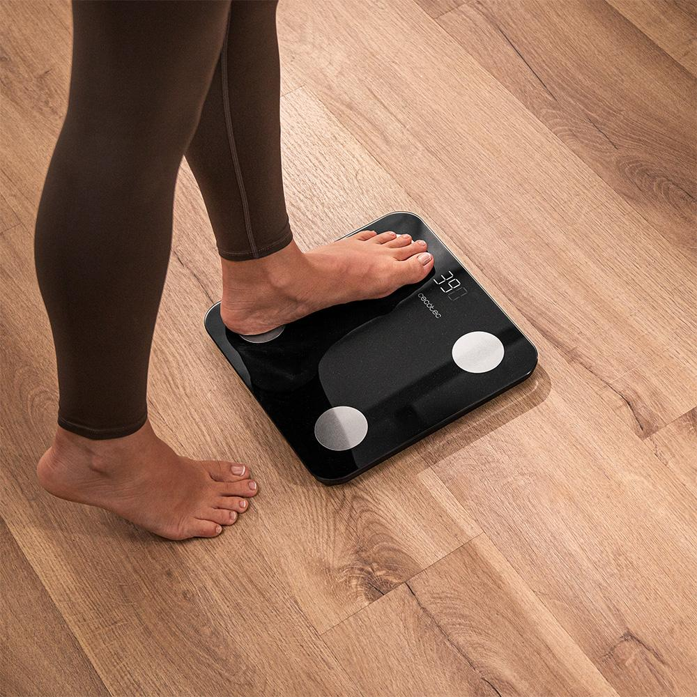 Una persona está de pie sobre una báscula digital negra. La pantalla muestra 39.0. El suelo es de tablones de madera. Se ven leggings marrones.