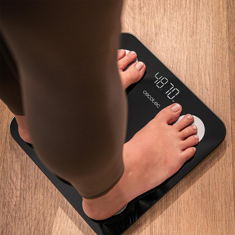 Una persona está de pie sobre una báscula digital negra. La pantalla muestra 48,70 kg. Se ven leggings marrones y suelo de madera.